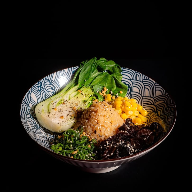 A colorful ramen bowl with bok choy, corn, rice, and dark seaweed on a black background.