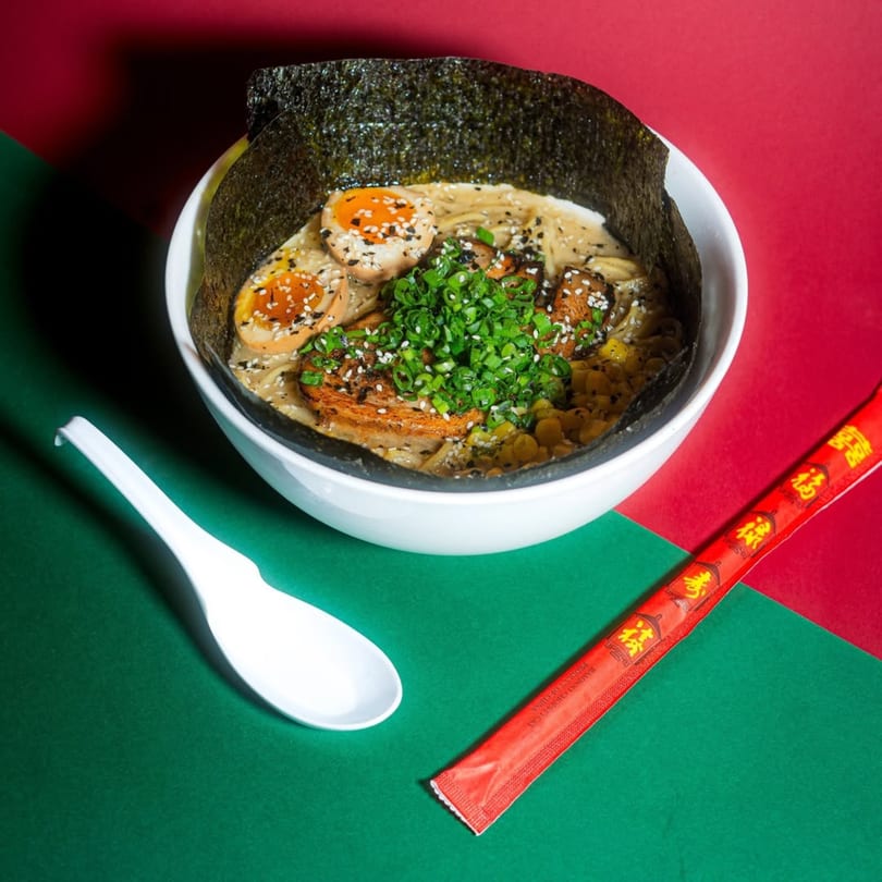 A bowl of Frankie Ramen with eggs, green onions, corn, and nori, with a spoon and chopsticks.