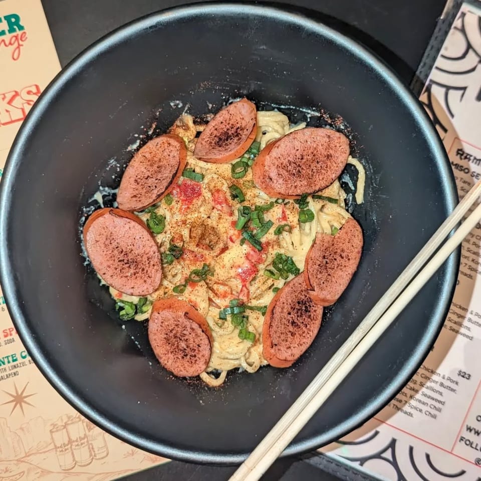 Frankie Ramen's creamy ramen with sliced sausage, green onions, and red spices in a black bowl.